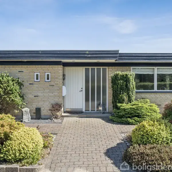 En gulstensbungalow med en hvid dør og store vinduer. Huset er omgivet af en velplejet have med buske og træer samt en brolagt sti, der leder til indgangen.