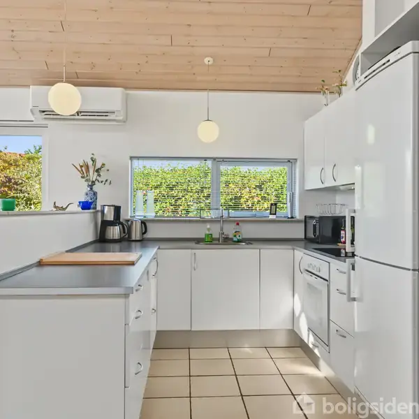 Et moderne køkken med hvide skabe og grå bordplader står i et lyst rum. Der er en elkedel, planter, en mikrobølgeovn og store vinduer med udsigt til en grøn have.