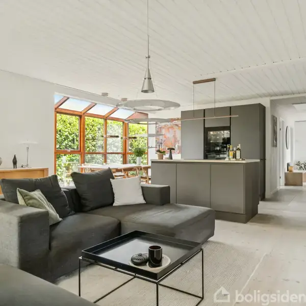Sofa i stueområde med grå farve og puder, placeret foran et lille sofabord. Omgivet af åbent køkken med mørke skabe og spiseplads ved store vinduer med udsigt til have.