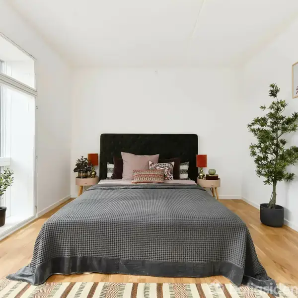 En seng med sorte og grå sengetæpper står i midten af et lyst soveværelse. Rummet har planter, hvide vægge, træmøbler og glasdør, der fører ud til balkon.