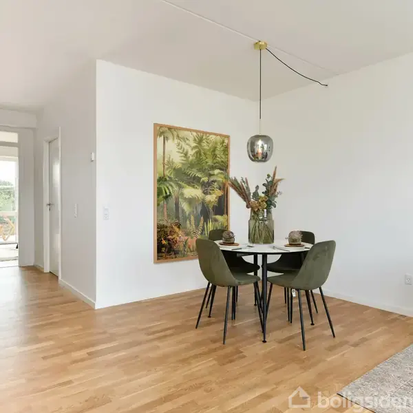 Et rundt spisebord med fire stole står i et lyst rum med trægulv. En dekorativ vase med blomster er placeret på bordet, og en malet væg hænger over det.