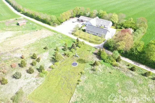 Gårdkompleks set fra luften omgivet af grønne marker. På græsplænen foran bygningerne er en trampolin placeret. Træer og en vej ses rundt om ejendommen.