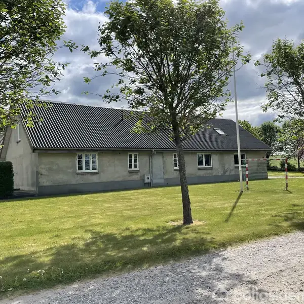 En grå bungalow med sort tag står på en velplejet græsplæne omkranset af træer. I forgrunden ses en grusvej og et flagstang. Skydet synger mellem grene.