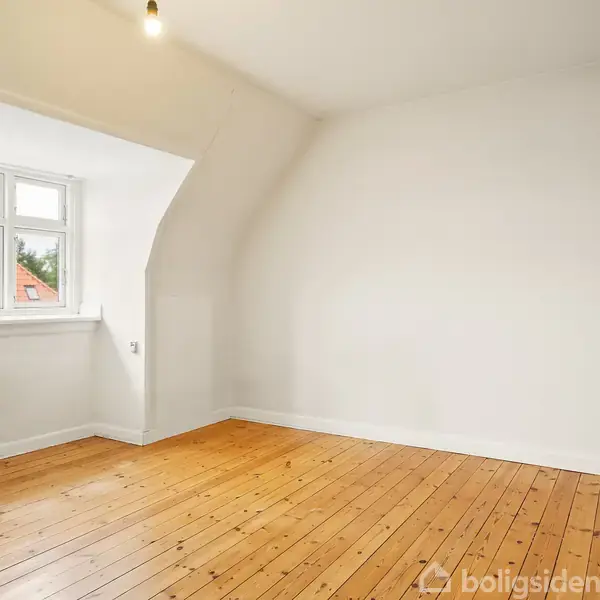 Et tomt rum med trægulv og hvide vægge har et vindue med udsigt til rødt tag. Radiator er placeret på højre væg. Ingen tekst fundet.