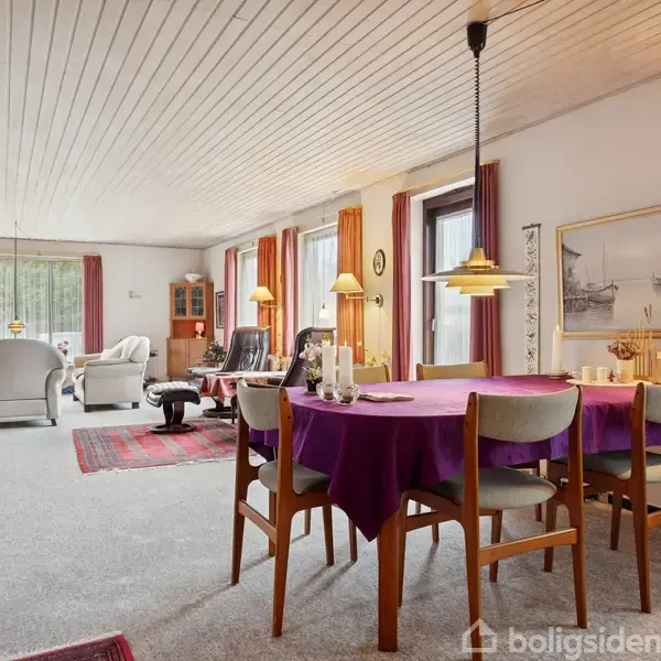 Et spisebord med lilla dug står omgivet af stole i en stue. Rummet har hvide vægge, flere vinduer med gardiner, sofaområde, hylder og hængende lamper.