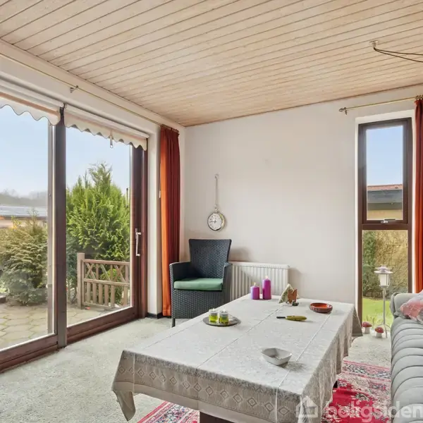 Stue med sofa og bord dekoreret med lille ur, beliggende foran store glasdøre med rødbrune gardiner, som åbner ud til terrasse med frodig have.