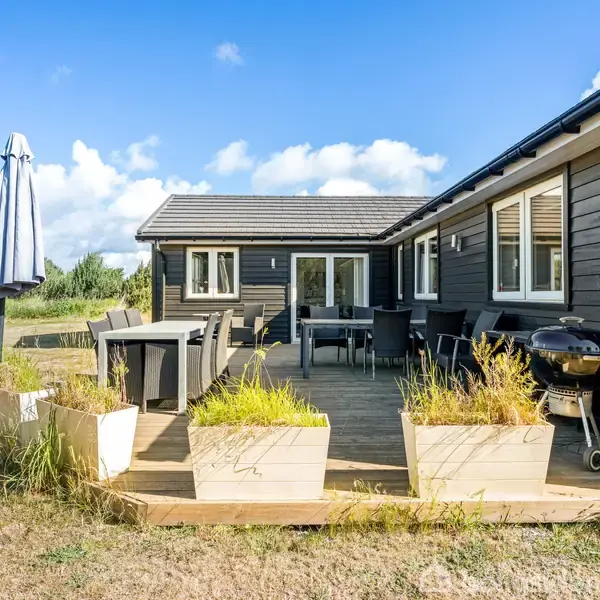 Sort sommerhus med terrasse og havemøbler, omgivet af græsplæne og træer i baggrunden. Terrassen har parasol, bord, stole og grill. Klare himmel og dagslys giver en lys atmosfære.