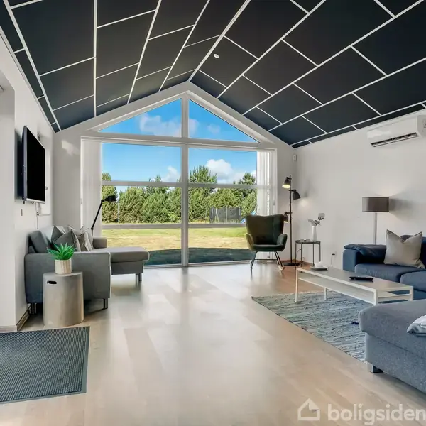 Stue med moderne interiør; en stor, grå sofa står til højre, mod et stort vindue der viser en have. Der er et sort-hvidt loft, og stuen har lyse trægulve.