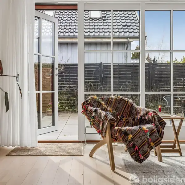 En stol dækket af et farverigt tæppe står ved siden af et lille bord. Døren står åben mod en terrasse med fliser og hegn. En plante står i hjørnet.