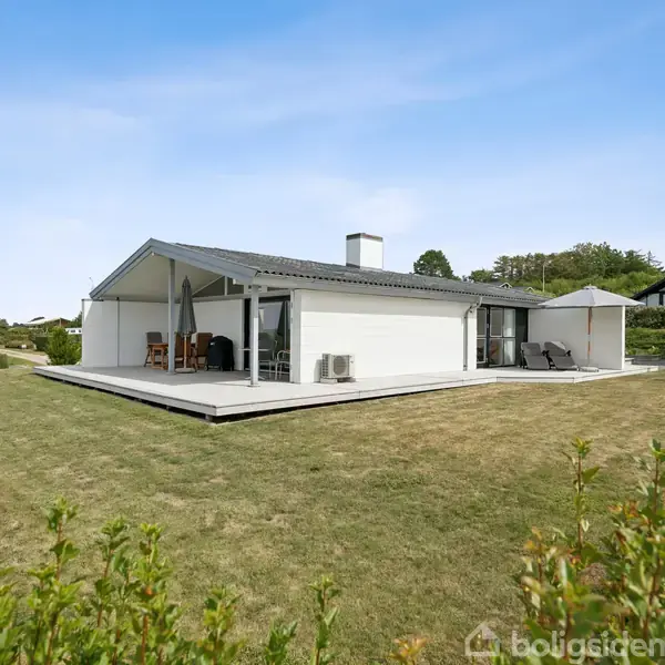 En moderne, hvid villa står stille på en velplejet græsplæne med en stor terrasse omgivet af buske og træer i et roligt boligområde.