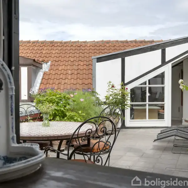 En gårdhave med jernmøbler og blomster i potter, omgivet af en bygning med hvid facade og rødt tegltag, skaber en hyggelig atmosfære under en overskyet himmel.