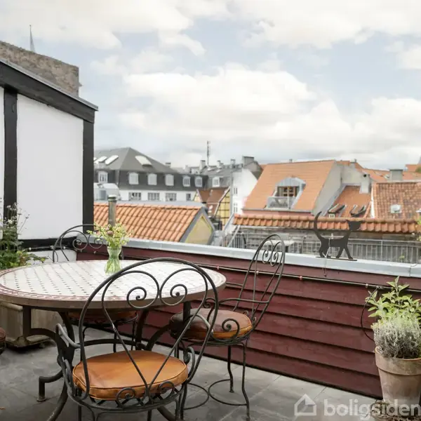 Rundt havebord med jernstole står placeret på en altan. Bordet har en vase med blomster. Omgivelserne består af hustage og bygninger, der danner en hyggelig, urban atmosfære.