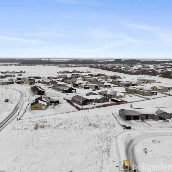 Boligområde dækket af sne set fra oven, hvor huse står tæt og veje forbinder dem. Landskabet er fladt og vidstrakt med flere bebyggelser i horisonten.