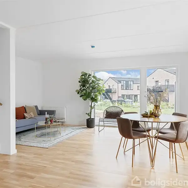 Stue med trægulv og store vinduer. En rund spisebord med fire stole står til højre, en sofa og et stuebord til venstre. En plante ved vinduet.