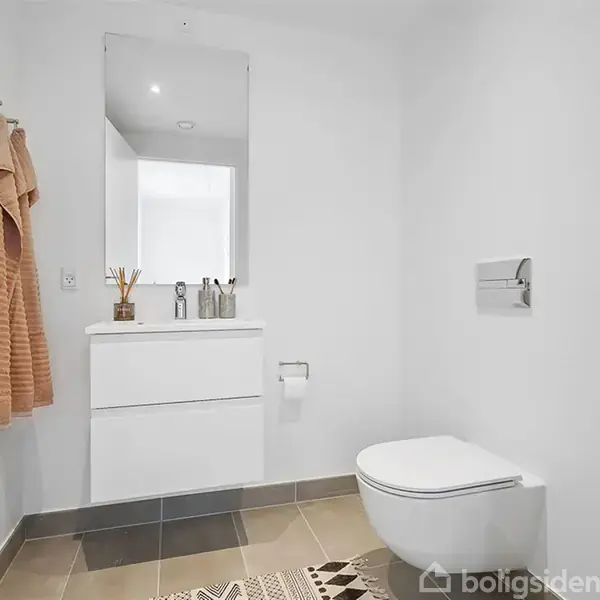 Et moderne badeværelse med flydende toilet og hvidt skab under spejl. Håndklæder hænger på en krog ved døren, og rummet har brune fliser på gulvet.