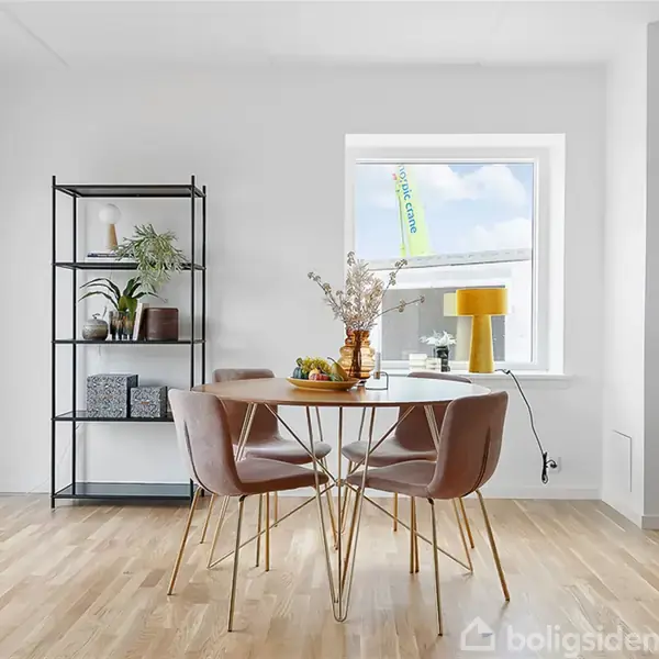 Et rundt bord med fire rosa stole står i et lyst rum ved et vindue. En åben reol med dekorationer står ved siden af. Laminatgulv bidrager til den moderne indretning.