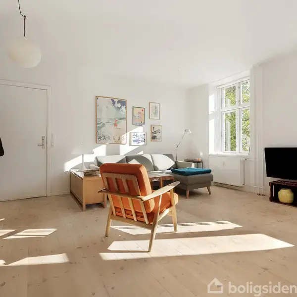 En lys stue med en orange lænestol foran en grå sofa, omgivet af billeder på væggen. Et fjernsyn står ved vinduet, og der er trægulv og bogreol.