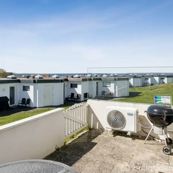 Græsplæne foran række af hvide bygninger ses fra en terrasse med grill og ventilationsanlæg. Et skilt viser teksten "Til salg". Havudsigt i baggrunden under en klar blå himmel.