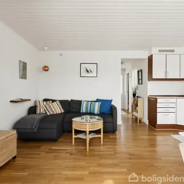 Stue med mørkegrå sofa fyldt med stribede puder står langs en hvid væg. Et lille rundt bord foran sofaen. Køkken til højre med hvide skabsdøre. Verdenskort på væggen.