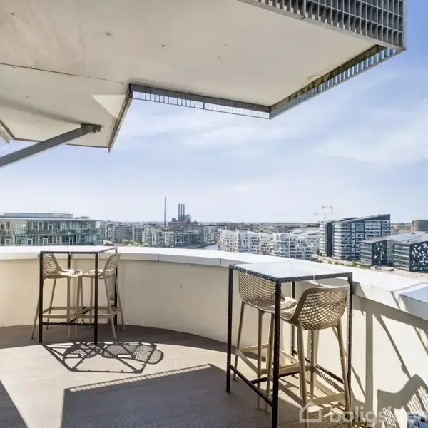 En balkon med høje barstole og et bord, stående stille under et halvtag, med udsigt over en moderne by med højhuse og klart blå himmel.