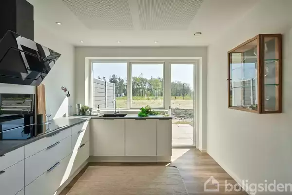 Et moderne køkken med hvide skabe og sort kogeplade ligger ved siden af en udgang til en have. Store vinduer giver udsigt til grønne træer.