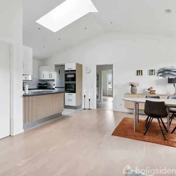 Et lyst køkkenalrum har en moderne køkkenø med trælameller og spisebord med to stole på et orange tæppe. Hvide vægge og loft med ovenlys skaber en luftig atmosfære.