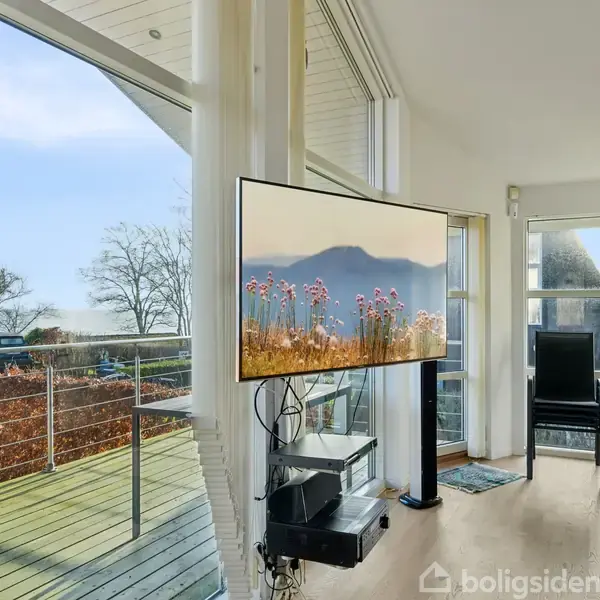 Fladskærms-TV viser bjerglandskab med blomster, ophængt i en moderne stue med store vinduer, som giver udsigt til en terrasse og have med træer.