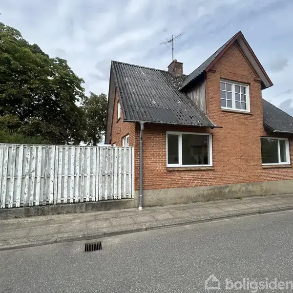 En rød murstensvilla med sort tag står ved siden af en hvid træhegn. Huset er placeret på en stille vej med træer i baggrunden.