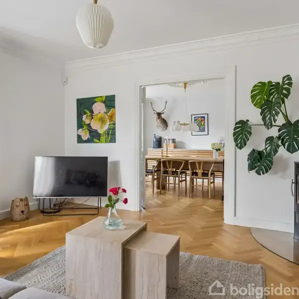 En moderne stue med en sofa og sofabord i front. TV'et står til venstre, og en pejs står til højre ved siden af en stor grøn plante. Bagved ses en spisestue med et stort bord.