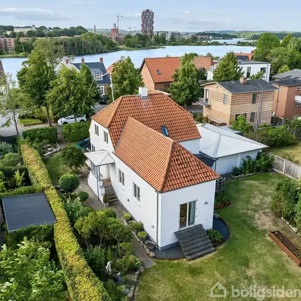 Et hvidt hus med rødt tag, omgivet af have og træer, ligger i et beboelseskvarter nær en sø. Baggrund viser flere huse og træer langs vandet.