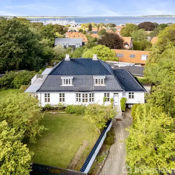 Et stort, hvidt hus med sort tag står omkranset af en frodig have. Det ligger i en villakvarter med flere huse og træer, og i baggrunden ses havet og en by.