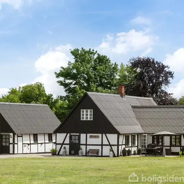Bindingsværkshus med stråtag og hvid facade står stille i en grøn have omgivet af træer. Sollys skaber en hyggelig atmosfære under den blå himmel med skyer.