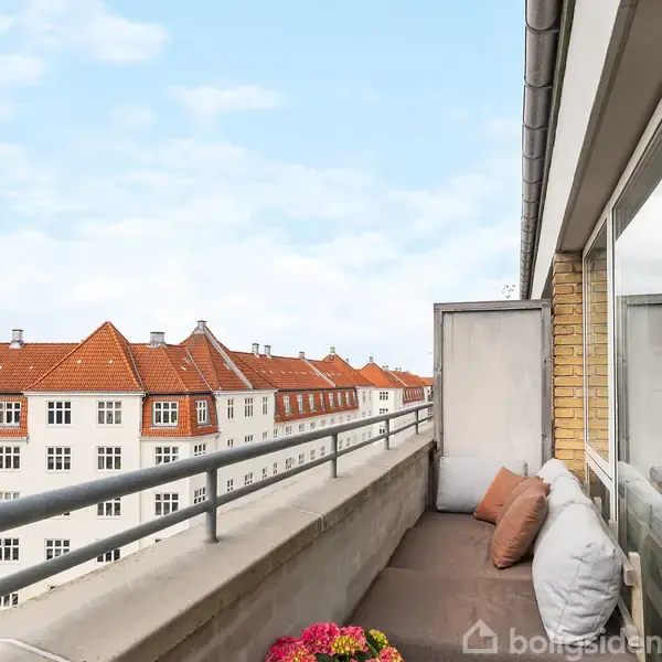 Altanen med rækværk rummer en lille sofa med puder og tilbyder udsigt til hvide bygninger med røde tage under en blå, skyfri himmel.