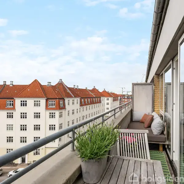 Balkon med træbord og planter. Stole er placeret omkring bordet. Baghusene i røde tage og hvide facader udgør baggrunden i en byscene under en delvist skyet himmel.