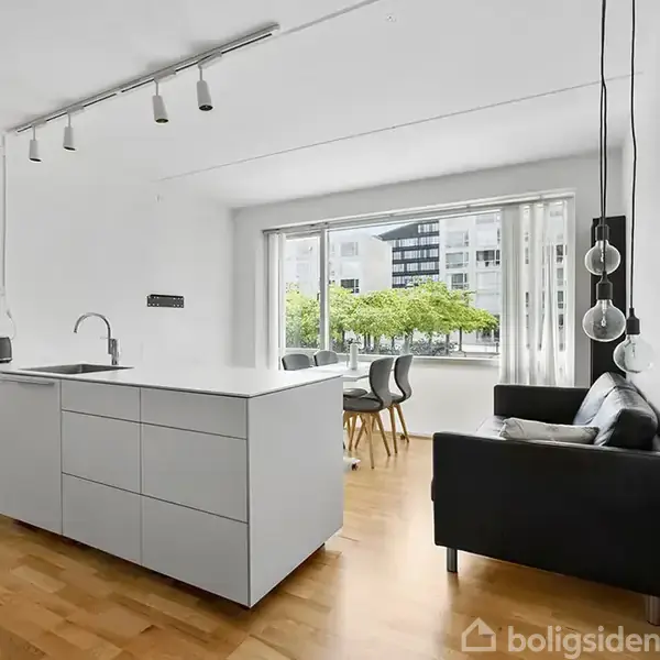 Et lyst køkkenalrum med en moderne køkkenø, hvor en sort sofa og spisebord står i et åbent rum. Store vinduer giver udsigt til træer og udendørs bygninger.