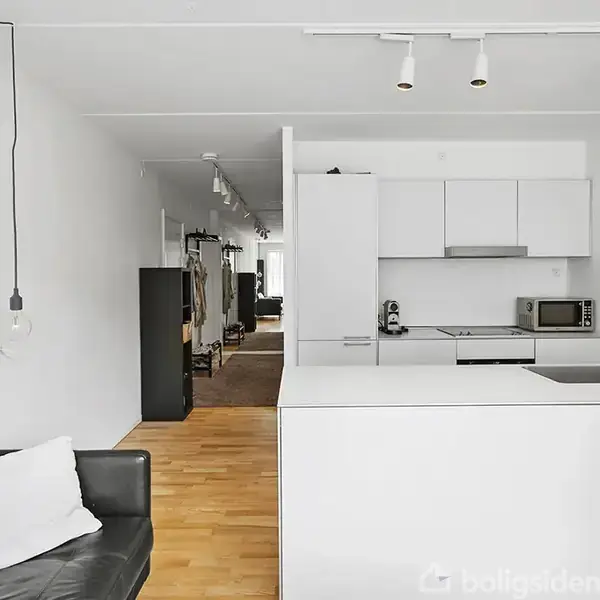 Sort lædersofa med hvid pude står i moderne køkken med hvide skabe; hvide hængelamper hænger ned. Trægulv strækker sig mod en gang med garderobe.