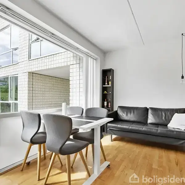 Spisebord med fire stole står ved en stor glasdør, der åbner ud mod en altan. En grå sofa og en smal reol ses i baggrunden.