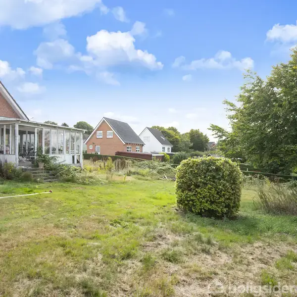 Hus med rød murstensfacade og tilbygget udestue. Stående i en have med græsplæne og buske. Naboer og træer ses i baggrunden under en blå himmel med skyer.