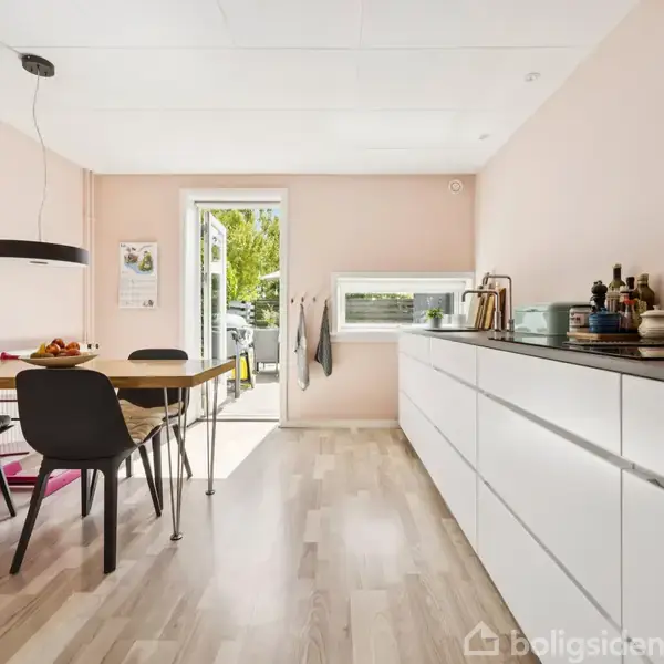 Køkkenbord står dækket med frugt i en moderne, lys spiseafdeling. Til venstre hænger en sort lampe over bordet, mens døren i baggrunden åbner ud til en grøn have.