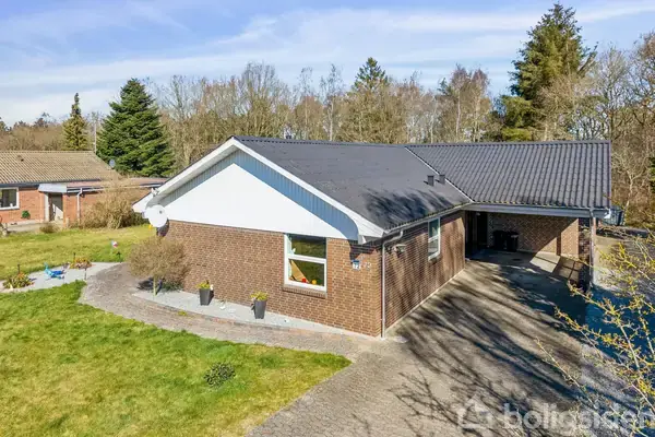 Et etplanshus med brun murstensfacade og sort tag ligger i en grøn have med græs og træer. Huset har en indkørsel og et tilstødende skur.