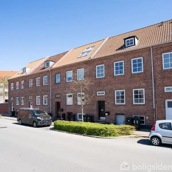 Rækkehuse af mursten står langs en stille vej med parkerede biler på en solrig dag. Husene har røde tegltage og hvide vinduer, og vejen er næsten tom.