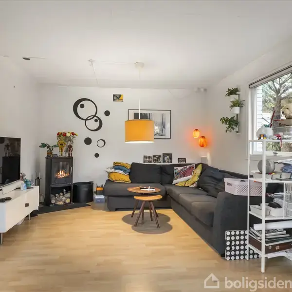 Stue med en stor grå sofa i hjørnet, hvor et rundt sofabord står foran. Pejs til venstre, orange hængelampe ovenover. Dekorative vægpynt. Vinduer til højre. Klokken på kommoden viser 9:44.