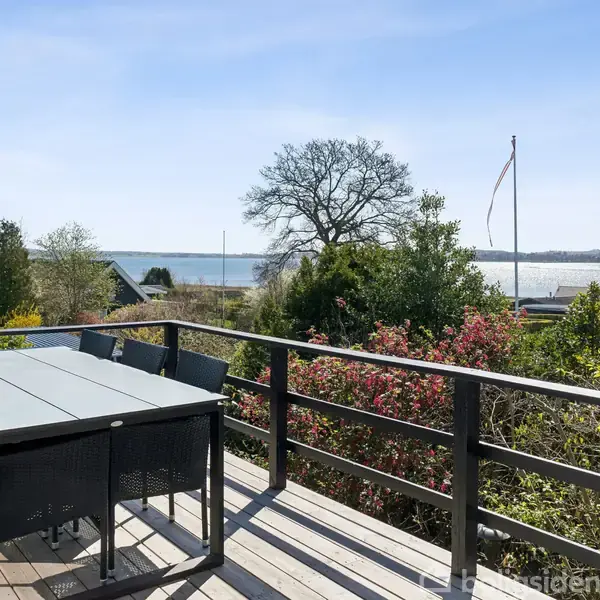 Terrassebord med seks stole står på en træterrasse. Udsigt over en have med farverige buske, et stort træ, og en fjord i baggrunden. Flagstang står i haven.