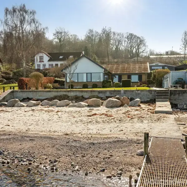 Hus ved kysten med glasfacade og rødt tag. Foran er en sandstrand og store sten. I baggrunden ses en skråning med træer og flere huse.