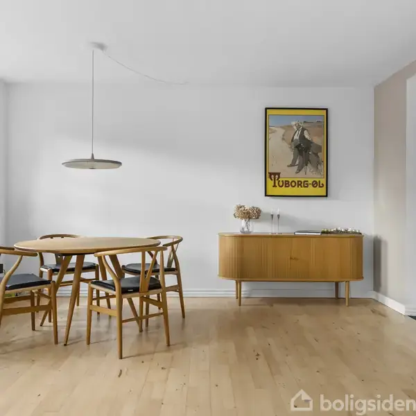 Et rundt spisebord med fire stole står i et lyst rum. På væggen hænger en plakat med teksten "Tuborg-Øl". Et skab står ved siden af en stor åbning til køkkenet.