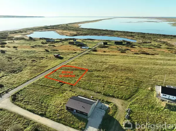 Grønt landareal med teksten "Fritidshus grund" skrevet inden for en rød markering. Nærliggende huse og veje ses, med en sø og kystlinje i baggrunden under en klar himmel.