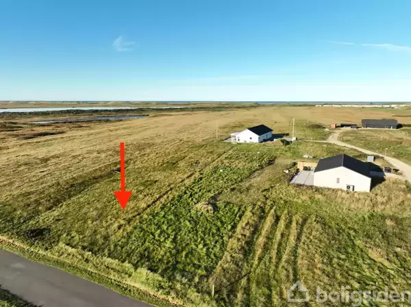 En rød pil peger mod en græsklædt grund ved siden af to ensomme bygninger på en åben landstrækning. En landevej løber gennem det grønne landskab under en klar blå himmel.