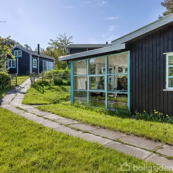 Mørkt træhus med hvide vinduesrammer står i sollys. Brostensti fører til huset gennem frodig, grøn have. Enkelt hus i baggrunden.
