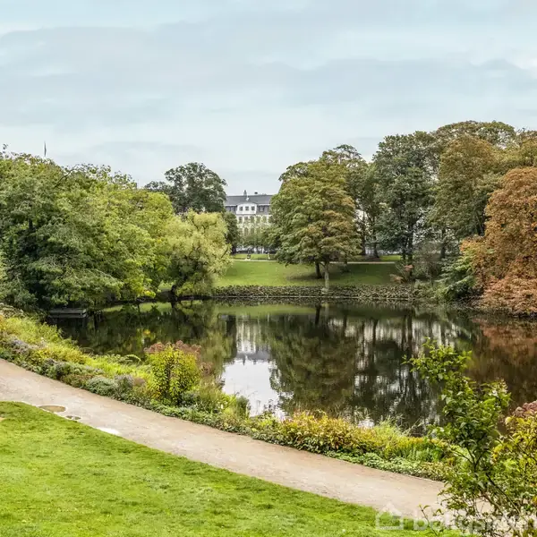 En sø reflekterer omgivelserne midt i en park, omkranset af grønne og efterårsfarvede træer. En sti løber langs søen med en statue nær venstre kant.