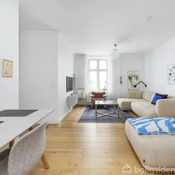 Stue med en lys sofa og sofabord i midten, fladskærms-TV på væggen. Arbejdsområde med skrivebord og skærm til venstre. Store vinduer giver naturligt lys. Malerier og plakater pryder væggene.